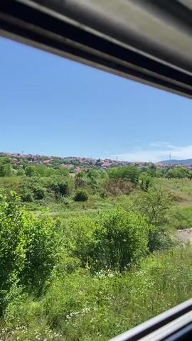 The video captures a moving shot from the perspective of someone inside a vehicle. The view is of a lush green landscape with a clear blue sky. In the foreground, there are dense trees and foliage. In the background, a town with red-tiled roofs is visible on a gentle slope, surrounded by more greenery. The sun is shining brightly, indicating a clear day. The overall impression is of a scenic journey through a verdant, semi-urban area.