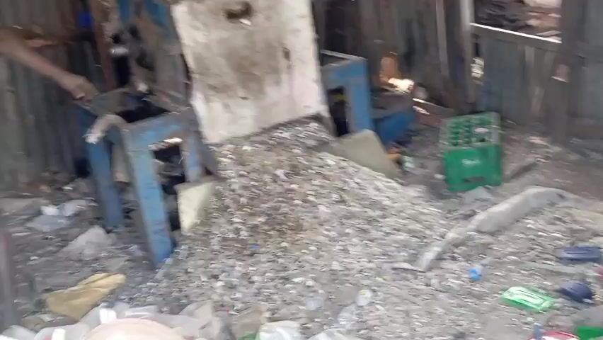 The video shows the inside of a cluttered, possibly industrial space. A large blue machine, possibly a shredder, stands prominently on the left. A pile of shredded material, appearing silver or metallic, accumulates beneath it. Debris litters the floor, including plastic bags, containers, and a green plastic crate. The walls are made of corrugated metal, and the space seems dimly lit. One person, wearing light-colored clothing, is visible on the left side of the frame. The machine appears to be in operation, as a loud noise is heard.