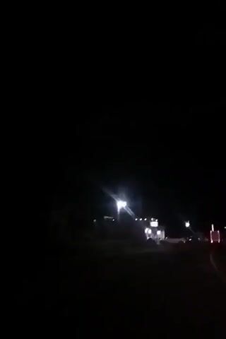The video shows a dark scene at night. A road is visible in the foreground. In the distance, there are buildings and vehicles with lights on. A large truck with its rear lights illuminated is parked. Another truck passes by. The overall visibility is low due to the darkness.