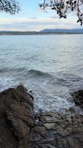 The video shows a rocky shoreline with the ocean in the foreground and a distant town on the horizon. The water is blue and there are some small waves breaking on the rocks. The sky is cloudy and the sun is setting. There are pine trees in the upper part of the frame. There are no people visible in the video.