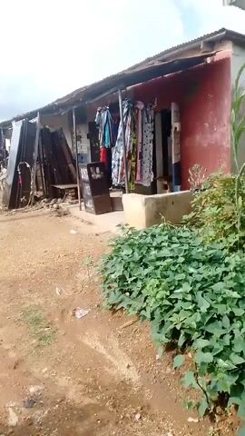 The video shows an outdoor scene. In the foreground, there is a dirt path with some green vegetation on the right side and scattered litter. In the middle ground, a simple storefront is visible, with clothes hanging outside for display. A dark-colored display stand is placed in front of the store. To the left of the store, there are metal objects and materials stacked up. In the background, two men are seated at a table, with one man standing. There are more metal structures and what appears to be a street or road with a sign in the distance. The sky is overcast.