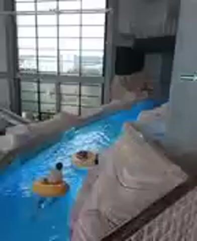 The video shows an indoor water park with a lazy river. Several people are floating on inner tubes in the bright blue water. The lazy river winds through a tropical-themed environment, with artificial rock formations and lush greenery. In the background, there are water slides and other attractions. The overall atmosphere appears to be fun and relaxing. The lighting suggests it's daytime, possibly a bright day given the reflections in the water, and the indoor setting means weather is not a factor. There is a green sign with an arrow on a grey pillar, but the text is not legible. The video appears to be taken at a water park in Reda, Poland.