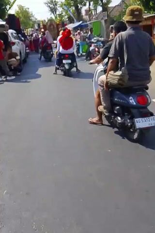 The video shows a street scene with several people and vehicles. In the foreground, a dark colored car is parked on the left side of the road with its trunk open. Several people are gathered around the car, some standing and some crouching. A person in a red shirt is visible near the open trunk. Further down the street, there are several motorcycles and some people walking. A few stalls or small shops are set up on the right side of the road. The scene appears to be outdoors on a sunny day, with trees and buildings lining the street. A procession or gathering of some sort is suggested by the presence of decorated elements in the background and the concentration of people.