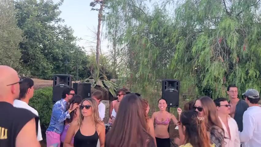 The video shows a group of young people at an outdoor gathering. In the foreground, a bald man wearing a black t-shirt with text is partially visible from the back. A woman with long dark hair in a black tank top is looking to her left. To her right, a woman with long blonde hair and sunglasses is also looking left. Behind them, a group of young men and women are interacting. Two large speakers are positioned on the ground on either side of a tall, thin tree with greenery. Several people are dancing or standing in conversation. One woman in a bikini top is smiling and looking forward. The environment appears to be a garden or wooded area with dense foliage in the background. The lighting suggests it is daytime, likely late afternoon or early evening.