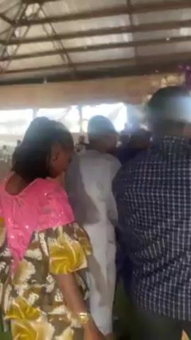 The video shows a wedding reception area with people dancing near a floral archway. The camera pans to the side, showing a woman walking and carrying a plastic bag. The area has green turf on the floor and appears to be in a tented structure. Decorations are visible in the background.