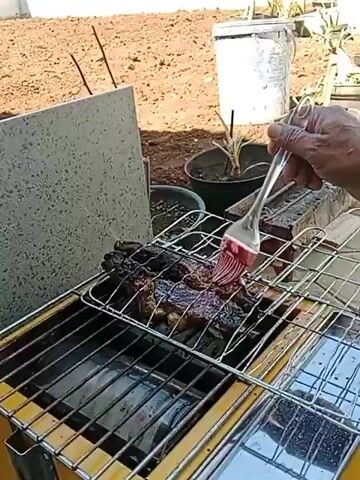 The video shows a person's hands holding a brush and applying a red sauce to meat that is cooking on a grill. The grill appears to be a portable one, possibly a gas grill, with a yellow frame and metal grates. The meat is dark brown and appears to be well-cooked or grilled. The background is an outdoor scene with dry, reddish-brown soil, some potted plants, and a concrete block structure. The lighting suggests it is daytime.