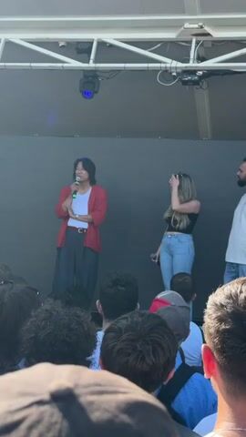 Three people stand on a stage in front of a dark backdrop. A person in the center, wearing a red cardigan over a white t-shirt and dark pants, holds a microphone and appears to be speaking. To their right, a woman in a black tank top and blue jeans also holds a microphone and touches her hair. To the far right, a man in a white t-shirt and blue jeans stands looking forward. The stage is covered by a white canopy structure with lighting fixtures visible. The scene is outdoors, likely during the daytime due to the bright lighting. A crowd of people is visible in the foreground, looking towards the stage. The event appears to be a public speaking engagement or a panel discussion, possibly related to the "Fast and Furious" franchise as suggested by the audio. The overall atmosphere seems to be casual and engaging. The location is identified as Cesena, Italy.