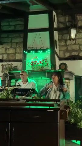 A man and a woman are performing on a stage. The man is seated behind a keyboard, wearing a white shirt. The woman is standing and singing into a microphone, wearing a denim jacket over a t-shirt. They are in a dimly lit room with a stone fireplace in the background. There are decorative lights and plants around the stage. The overall atmosphere appears to be a live performance in a restaurant or bar.