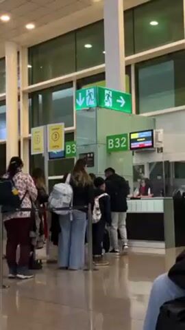 In an indoor airport scene, a group of people are queuing at gate B32. The people are of various ages, including children, and are wearing casual clothing and backpacks. An airport employee wearing a red scarf sits behind a counter, seemingly checking boarding passes or documents. Above the counter are illuminated signs indicating the gate numbers and an exit sign with arrows pointing down and to the right. The airport environment appears modern, with large windows in the background. The lighting is bright, and the overall scene suggests the typical hustle and bustle of an airport.