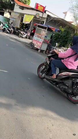 A woman wearing a blue hijab and a pink long-sleeved shirt rides a black motorcycle down a paved road. She is wearing blue jeans. The motorcycle has red accents on its wheels. In the background, there are food stalls and several parked motorcycles. The weather appears to be sunny.