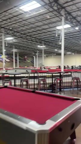 The video shows the interior of what appears to be a large entertainment venue. The foreground contains a pool table with a red felt surface. Beyond the pool table is a seating area with high tables and stools, delineated by a railing. In the background, a go-kart track is visible with several go-karts on the track. The building has a high ceiling with exposed metal beams and support pillars, and skylights are present. The floor appears to be polished concrete. There are no people clearly visible in the video.