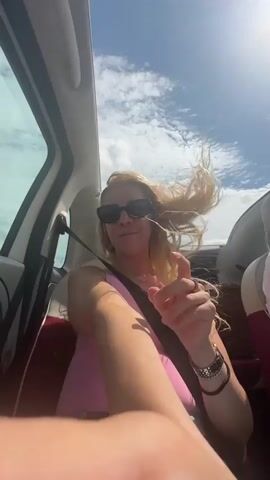 The video is a first-person perspective shot from inside a car. A woman with blonde hair and sunglasses is visible in the passenger seat, wearing a pink top and a seatbelt. Her hair is being blown back by the wind. She smiles and makes a face at the camera, pointing. The car appears to be on a highway with a cloudy sky and distant trees. Luggage is visible in the back seat.