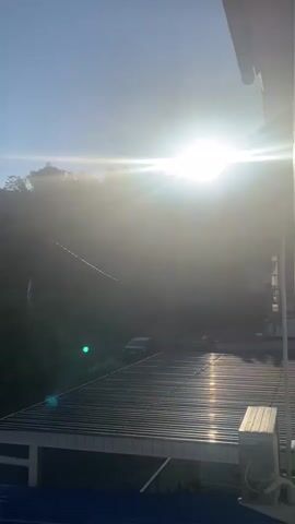 The video shows a bright sun shining in a clear blue sky. The sun is positioned in the upper right quadrant of the frame, creating a lens flare that obscures some of the details in the scene. In the foreground, there is a dark, corrugated roof, possibly part of a building. To the left of the roof, a white railing is visible, and a small, light-colored object, perhaps an air conditioning unit, is on the roof. Beyond the roof, a lush green hillside is visible, covered in trees. Several power lines stretch across the hillside, and a utility pole is also present. A small white flag is hanging from a line on the hillside. A dark-colored car is parked on a road that winds up the hill. The overall scene is bathed in bright sunlight, with the sun's rays creating a hazy glow.