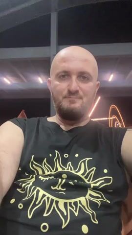 A man with a shaved head and a beard smiles at the camera. He is wearing a black t-shirt with a gold sun design on it. The background appears to be outdoors at night, with some lights visible.