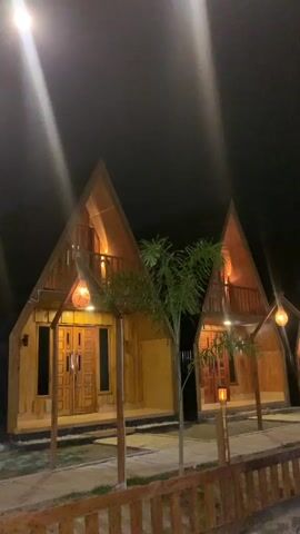 The video captures a night-time scene featuring a row of several wooden cabins, likely part of a resort or lodging area. The cabins have distinctive pointed roofs and small balconies, with warm yellow lights from wall sconces and hanging lanterns illuminating their wooden facades and the pathways in front of them. Small, sparse trees are planted between the cabins. A low wooden fence runs along the bottom of the frame, separating the viewer from the structures. The sky is dark, indicating night, with streaks of bright light visible in the upper part of the frame, possibly from distant light sources or reflections. The camera slowly pans from left to right, showcasing the sequence of cabins. No people or text are visible.