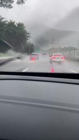 The video, captured from inside a vehicle, shows a highway in rainy conditions. Several cars are visible ahead on the wet road, their taillights illuminated. The visibility is reduced due to the rain and mist, with mountains visible in the background obscured by clouds. Palm trees line the left side of the highway, and a guardrail is present on the right. Water is splashing up from the tires of the vehicles, indicating heavy rain and potentially standing water on the road. The windshield of the vehicle from which the video is taken is speckled with raindrops.