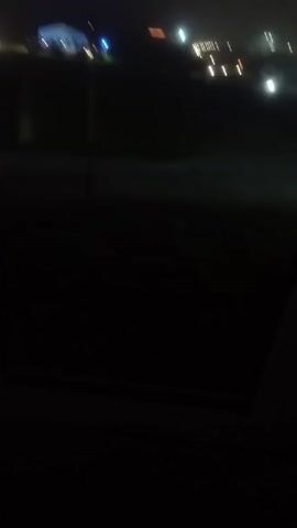 This video is a very dark, blurry, and shaky recording of an outdoor scene at night. There are some faint lights visible in the distance, possibly from a city or a road. The foreground is mostly black, with a hint of textured ground. The overall impression is of a low-quality recording made with a moving camera in poor lighting conditions. No people or specific objects can be clearly identified.