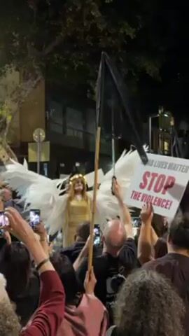 The video shows a crowd of people at night, possibly a demonstration or protest. In the center, someone dressed as an angel with large white wings and a gold headdress stands out. People in the crowd hold up their phones to record the scene. Some are holding signs, including one that reads "ARAB LIVES MATTER" and "STOP THE VIOLENCE!". Others are carrying black flags attached to wooden poles. The location appears to be an urban street lined with buildings. The atmosphere seems charged with emotion, as people are actively engaged in the event.