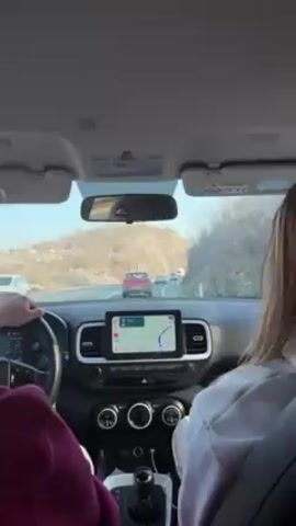 Inside a car, a person in a maroon sweater drives on a paved road with mountains in the background, while a passenger with blonde hair sits beside them. The car is equipped with a navigation system showing a blue route on a map. Other vehicles are visible ahead on the road, including a white van and red car. The scene is captured from the perspective of someone in the back seat, looking forward.