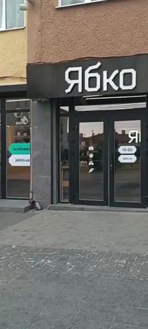 The video shows a series of urban scenes. It begins with a storefront that has Cyrillic text on its awning, likely a retail establishment. Next, the camera pans to another storefront, also featuring Cyrillic text, which appears to be a computer store. Then, the view shifts to a tree-lined pedestrian walkway with benches, leading to a pharmacy with a green cross symbol. Finally, the video shows a town square with buildings in the background and a prominent clock tower, suggesting a historical or administrative center. The overall impression is of a European town or city.