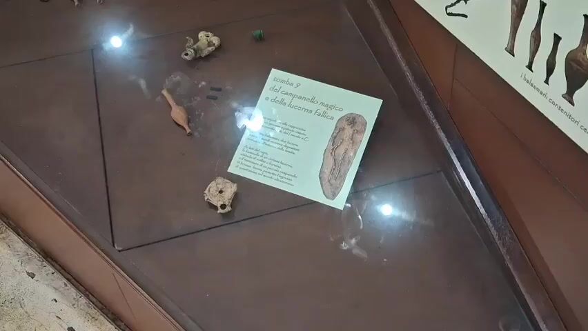 The video shows a museum display case with archaeological artifacts. The display case has a brown surface and is illuminated by overhead lights that cause reflections. On the surface are several small, ancient-looking objects, some appearing to be pottery fragments or small tools. A prominent item is a white informational card with Italian text and an image of what appears to be a phallic-shaped object. The text on the card mentions "tomba 9" and "campanello magico e della lucerna fallica," suggesting it relates to tomb number 9 and a magic bell and phallic lamp. Several small pottery pieces, a green bead, and what looks like a small wooden tool are also visible. The lighting creates glare on the glass surface, obscuring some details. The overall environment appears to be an indoor museum exhibit.