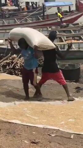 Two men are carrying a large white sack between them on their heads. One man wears a light blue shirt and maroon shorts. The other wears a black shirt and red patterned shorts. They are walking on a dirt surface covered with a layer of light-colored grains. Behind them are several boats and a pile of wood. In the background, water is visible.