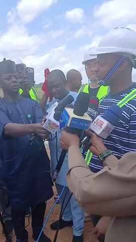 A group of men are gathered outdoors, possibly at a construction site, with a cloudy sky overhead. In the foreground, several microphones from different news organizations are being held up to a man in a blue traditional outfit and a black cap, who appears to be speaking. Behind him, other men, some wearing hard hats and high-visibility vests, are also present. One man in the center is holding his forehead, seemingly distressed or hot. Another man is holding a phone up, likely recording the event. The ground is rough and appears to be dirt or gravel. The overall scene suggests a press conference or interview.