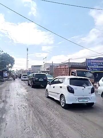 The video shows a view of a congested street with several cars and trucks. The road appears to be made of dirt and gravel, with some puddles of water. The sky is partly cloudy. In the foreground, a white car is parked on the right side of the road. Further down the street, there is a line of vehicles, including SUVs, vans, and trucks, moving slowly or stopped. On the left side of the road, there are some buildings and trees. There are also power lines and a tall communication tower in the background. The overall impression is of a busy, possibly rural or semi-urban area experiencing traffic congestion.