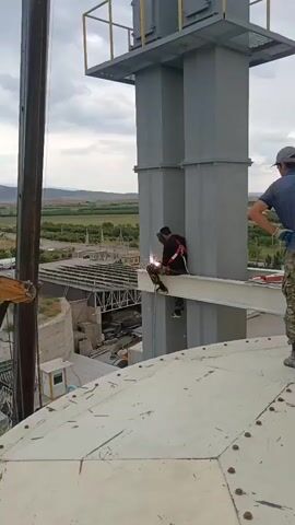 A construction worker is welding on a metal beam that extends from a tall, gray industrial structure. The worker is seated on the beam, wearing a safety harness, a welding mask, and work clothes. Sparks are flying from the welding torch. Another worker stands nearby, to the right of the frame, also wearing work clothes and a cap. In the background, a crane boom extends from the left side of the frame, and the main body of the crane is not visible. Further in the background, there is a sprawling industrial complex with buildings and a road, and beyond that, a landscape of rolling hills and vegetation under a cloudy sky. The scene appears to be an outdoor construction site at an industrial facility.