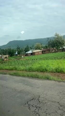 A video pans across a rural village scene. The foreground shows a cracked asphalt road with a grassy verge. Beyond the road, there are several simple houses with corrugated metal roofs and mud brick walls. Lush green vegetation, including crops and trees, surrounds the houses. In the background, rolling hills covered in trees form a backdrop under a cloudy sky. People are visible near some of the houses, engaged in what appear to be daily activities, and there are piles of wood or building materials. The overall impression is of a modest, agricultural community situated in a hilly, green landscape.