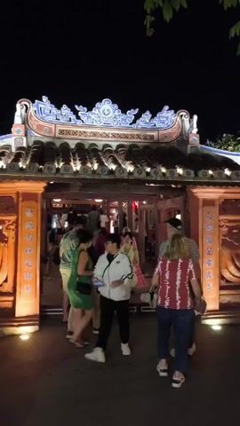 At night, several people pass through an ornately-decorated, glowing archway. The archway is topped with dark brown tiles and blue and white decorative elements.  The lighting is subdued, highlighting the structure. The people are dressed in a variety of casual clothing. They are walking in both directions through the archway. The scene appears to be in a historic or cultural area.
