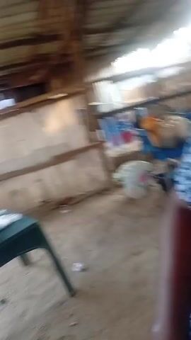 The camera pans around an outdoor structure with a corrugated metal roof. Inside, there's a green plastic table with a dark bag and water bottle on it. A woman in green leggings and a grey shirt sits on a green plastic chair at another table in the background. Outside the structure, there are other buildings and people. The ground is sandy.