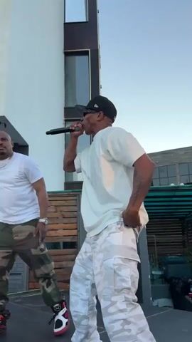 Two men are performing on a stage outdoors. The man in the foreground, on the right, is wearing a white t-shirt, a black baseball cap, sunglasses, and white camouflage pants. He is holding a microphone in his right hand and appears to be singing or rapping with his mouth open. His left hand is on his hip. The man in the background, on the left, is wearing a white t-shirt, a silver chain, and camouflage pants. He is also holding a microphone and looking towards the front. Both men are wearing sneakers. The setting appears to be a rooftop or elevated outdoor area with a building in the background. The sky is visible, suggesting it is daytime, possibly late afternoon or early evening given the lighting. Wooden elements and some greenery are visible in the background. There is some text on the building behind the man on the left, but it is illegible. The overall mood appears energetic and performative. The timestamp indicates the recording starts at 00:00.