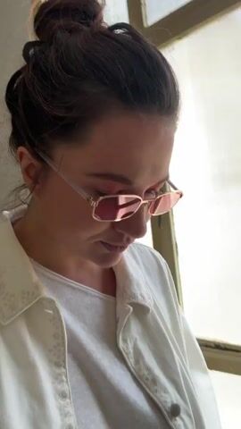 A young woman wearing rose-tinted rectangular sunglasses, a white t-shirt, and a white denim jacket stands indoors near a window with vertical blinds. Her dark hair is pulled back in a bun. She smiles and gives a slight kiss to the camera.