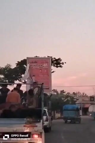 The video shows the back of a small white truck with a blue cargo bed driving down a road. Several young men are sitting and lying in the truck bed. A billboard stands by the side of the road, bearing a poster with the words “MILAD GAM 48” visible. A tall cellular tower is also visible. The time stamp on the video reads “OPPO Reno8 2026.05.23 06:17”. Other vehicles and pedestrians are also present. The sky is pale with the light of either sunrise or sunset.