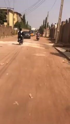 The video shows a street view in a residential area. The road is made of dirt and sand, with patches of concrete. There are buildings on either side of the road, and power lines overhead. Several vehicles are moving down the road, including motorcycles, a car, and a three-wheeled vehicle. The sky is bright and clear.