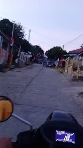 A first-person perspective video shows someone riding a motorcycle down a street lined with houses and trees. The sky is a pale blue, indicating either dawn or dusk. The road is paved but cracked, with some debris visible. A red and white flag, possibly the Indonesian flag, is displayed on one of the houses. Several cars are parked or driving on the street. The rider's hand is visible on the handlebars, and a yellow fabric is seen in the motorcycle's mirror. A digital display on the motorcycle shows the number 18, possibly indicating speed. The overall impression is of a casual ride through a residential area in the early morning or late evening.