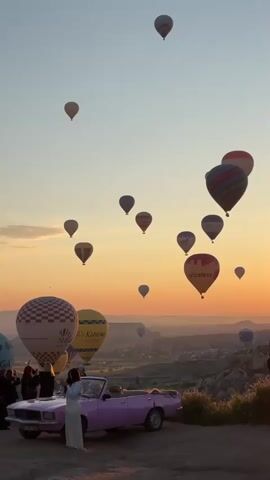 This image captures a breathtaking sunrise scene in Göreme, Cappadocia, Turkey, renowned for its hot air balloon flights. Numerous hot air balloons of various colors, patterns, and sizes ascend into a vibrant sky, transitioning from a warm golden-orange hue near the horizon to a soft blue overhead.

In the foreground, a light pink vintage convertible car is parked on an elevated dirt path, facing the scenic landscape. A woman in a long white dress stands beside the driver's side of the car, gazing towards the ascending balloons. Behind the car, a small group of people are also visible, observing the spectacular event.

The landscape features rolling hills and valleys characteristic of Cappadocia's unique geological formations, stretching into the hazy distance. Many balloons display visible text and logos; among them are "Sultan Ballons," "Air Cappadocia Ballons," "Royal Balloon," "Butterfly Balloons," "MEGA BALLOONS," and the "Priceless" logo associated with Mastercard.

The overall scene conveys a sense of wonder and tranquility, highlighting the iconic hot air balloon experience at dawn in this famous Turkish region. The sky is clear, and the soft, warm light of the rising sun bathes the entire setting.