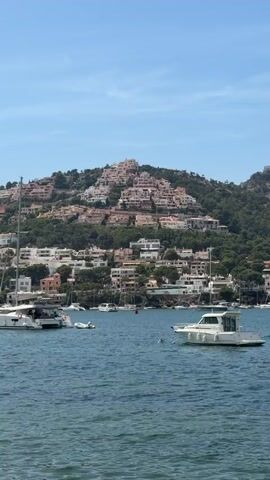 A daytime outdoor shot features a wide view of a coastal scene. The foreground shows calm blue water with several boats anchored. A large white catamaran is on the left, and a smaller white motorboat is on the right. In the middle distance, several other smaller boats are visible. The background is dominated by a lush green hillside dotted with numerous light-colored houses, forming a clustered settlement. The houses are built in terraces up the slope. The sky is clear and blue with no clouds. The overall impression is one of a serene and picturesque Mediterranean landscape.