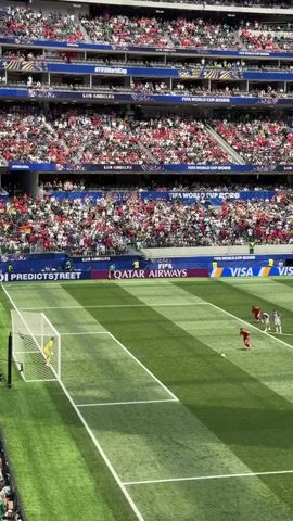 This is a high-angle video of a soccer game in a stadium filled with spectators. The field is green with white lines. In the foreground, several soccer players in red uniforms are running towards the camera, while two players in white uniforms are running away from the camera. A goalkeeper in a yellow uniform is positioned near the goal on the left side of the frame. There are advertisements visible around the perimeter of the field, including "FIFA WORLD CUP 2026," "QATAR AIRWAYS," and "VISA." The spectators in the stands are wearing a variety of clothing, mostly red and white. The action captured is a moment of play during the game.