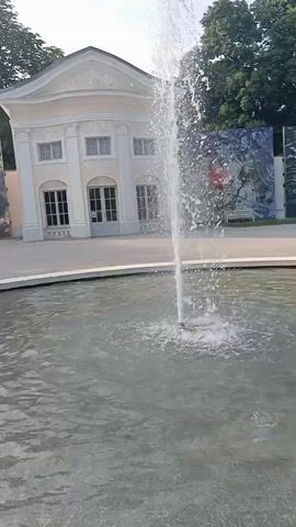 This media file shows a fountain in a body of water in front of a building with large, colorful artworks displayed on either side. The building is light yellow with a classical facade, featuring columns, arched windows, and a pediment. It appears to be a sunny day, likely in the late afternoon given the angle of the light and the shadows. The water in the foreground is rippling, with the fountain spraying upwards. The artworks on the walls are abstract and vibrant. Trees are visible behind the building. The overall scene suggests a pleasant outdoor setting, possibly a park or a public space associated with art and architecture. There is no visible text in the media file.