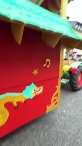 The video shows a vibrant parade with a tractor pulling a decorative float. The float has a red base with a green dragon and gold embellishments, and a green-tiled roof adorned with red lanterns. The side of the float features a sign for "HOFBRÄUNGER GARS/AM/INN" surrounded by cherry blossoms. People in colorful costumes are visible in the parade, including one individual dressed as a Japanese warrior. The float riders are handing out drinks to the crowd. The parade takes place on a street with buildings on either side and a gathering of spectators. The weather appears overcast.