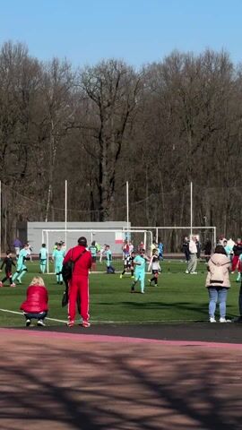 The video shows a youth soccer game in progress on a green field. The players are wearing light blue and black and white uniforms. Spectators stand along the edge of the field watching the game. A coach in a red tracksuit stands near the sideline. In the background, there is a row of bare trees and a clear blue sky. The game is focused near the goal.
