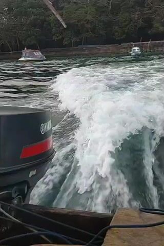 The video shows the wake from a boat engine. The engine is black with a red stripe and a white Yamaha logo. The water is choppy and white with foam. In the background, there is a line of trees and a smaller white boat. The sky is overcast. The perspective is from the back of the boat, looking out at the water.