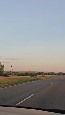 The video shows a view from inside a vehicle traveling on a paved road. The sky is clear and transitions from a pale blue at the top to a pale orange near the horizon. The road has a white line marking the center and a white line along the right edge. On the left side of the road, there are fields of grass and some trees. A tall wooden structure, possibly a power line tower or a windmill, is visible in the distance on the left. On the right side of the road, there are more trees and some vehicles parked along the roadside. The overall impression is a scenic drive through a rural landscape, possibly at sunset or sunrise due to the colors in the sky.