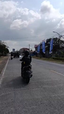 The video shows a view from a moving vehicle traveling on a road in what appears to be a tropical or subtropical region. The road is paved and has lane markings. On the left side of the road, there is a sidewalk, a dirt shoulder, and vegetation, including banana trees and other leafy plants. Power lines are visible overhead, supported by poles. Several blue flags with white text are displayed along the side of the road, some attached to poles. On the right side of the road, there are buildings that appear to be shops or houses, some with awnings.

In the foreground, on the left side of the road, a person wearing a helmet and dark clothing is riding a black scooter. The scooter has a license plate that is partially visible. The vehicle from which the video is filmed is traveling on the road, following the scooter and other vehicles.

Further down the road, other vehicles, including cars and motorcycles, are visible. The sky is partly cloudy with white clouds against a blue sky. The overall impression is of a sunny day in a populated area with roadside businesses.