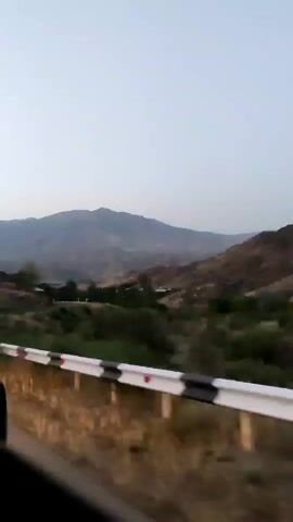 The video shows a landscape from a moving vehicle. The foreground features a roadside guardrail with white and red reflective markers. Beyond the guardrail, there is dry vegetation and a line of green trees. In the middle ground, rolling hills and mountains are visible under a clear sky. The lighting suggests it is either dawn or dusk.