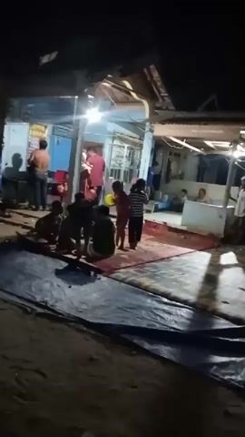 A group of people are gathered in an outdoor area at night. There is a small building with lights on, possibly a shop or house. Several individuals are standing and sitting, some are crouched. One person is wearing a red garment. A large blue tarp is spread on the ground in the foreground. The scene appears to be a social gathering.