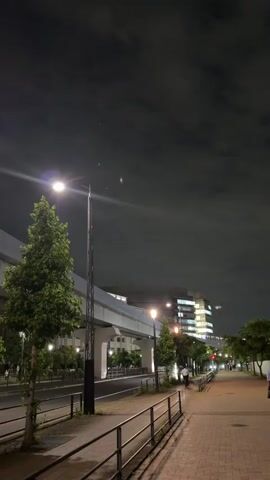 A nighttime street view shows a pedestrian walkway on the right side and a road with light traffic on the left. A raised pedestrian bridge or elevated train track spans the upper portion of the frame. Streetlights illuminate the walkway and road, casting long shadows. Trees line the sidewalk and the median separating the road. Buildings with illuminated windows are visible in the background. Several cars pass by on the road, and a few indistinct figures can be seen walking on the sidewalk. The sky is dark with scattered clouds.