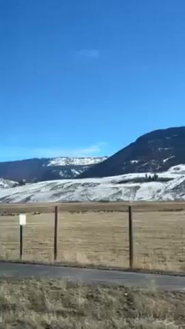 The video shows a view from a moving vehicle, likely a car, driving along a road. A field stretches out to the right of the road, fenced off with wooden posts. In the distance, mountains are visible, partially covered in snow. The sky is a clear, bright blue. There are road signs on the side of the road. The overall impression is that of a winter landscape.